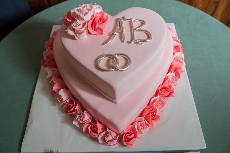 a pink heart shaped cake with roses and two initials on top, sitting on a table