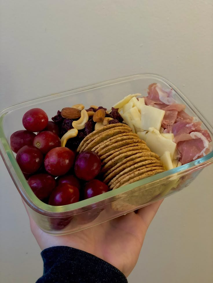 a hand holding a glass bowl filled with grapes, crackers and cheese next to nuts