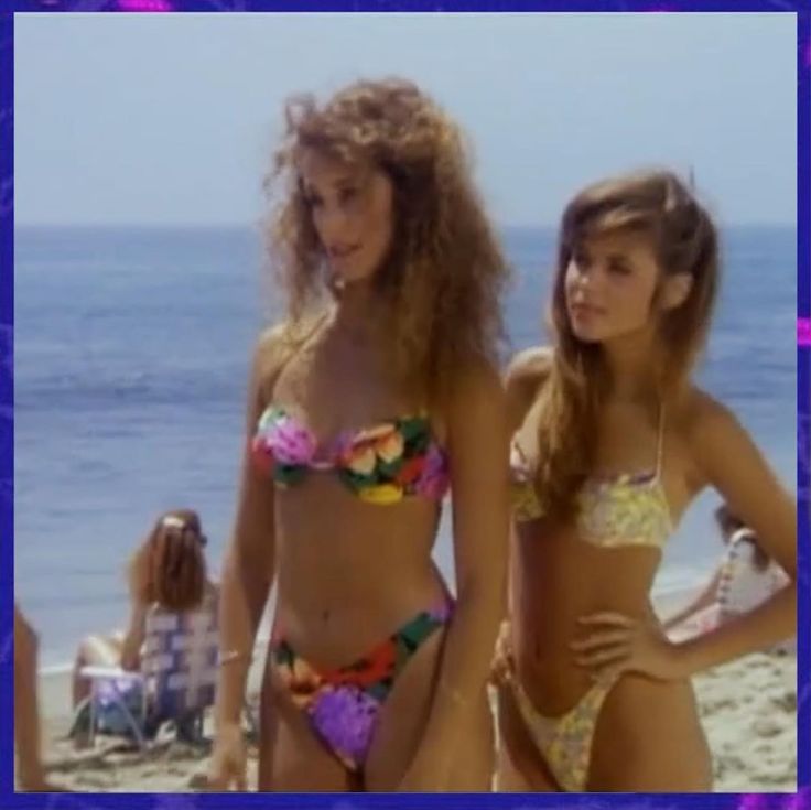 two women in bikinis standing on the beach next to each other and looking at the camera