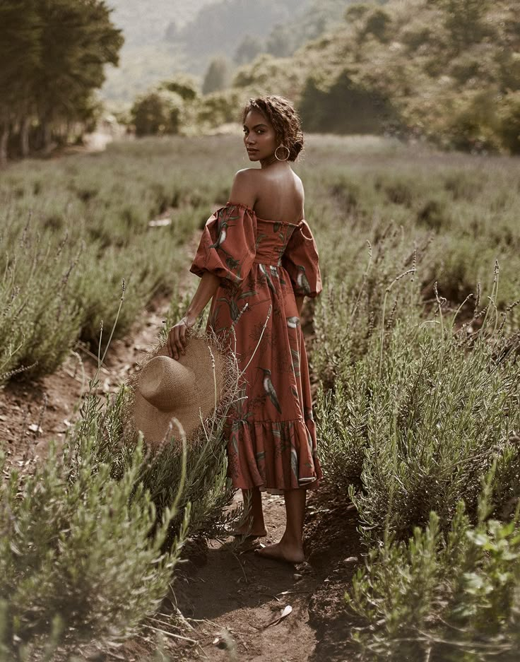 a woman standing in a field with a hat on her head and wearing a red dress