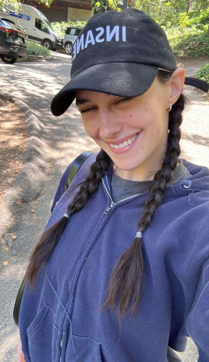 a woman with braids wearing a hat and smiling at the camera while standing in front of a tree