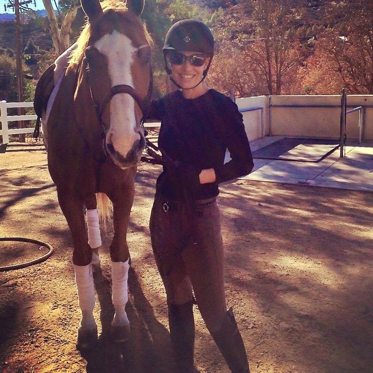 a woman standing next to a brown and white horse on dirt ground with trees in the background