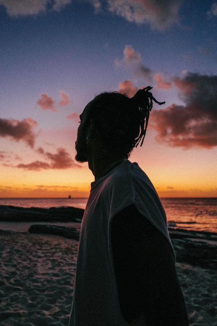 a man standing on top of a sandy beach next to the ocean at sunset with clouds in the sky