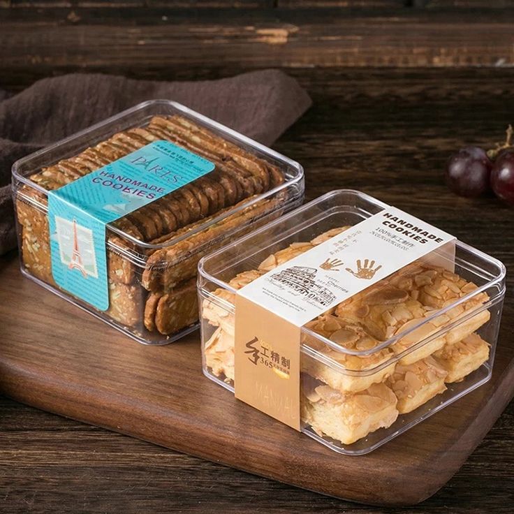 two plastic containers filled with crackers on top of a wooden cutting board next to grapes