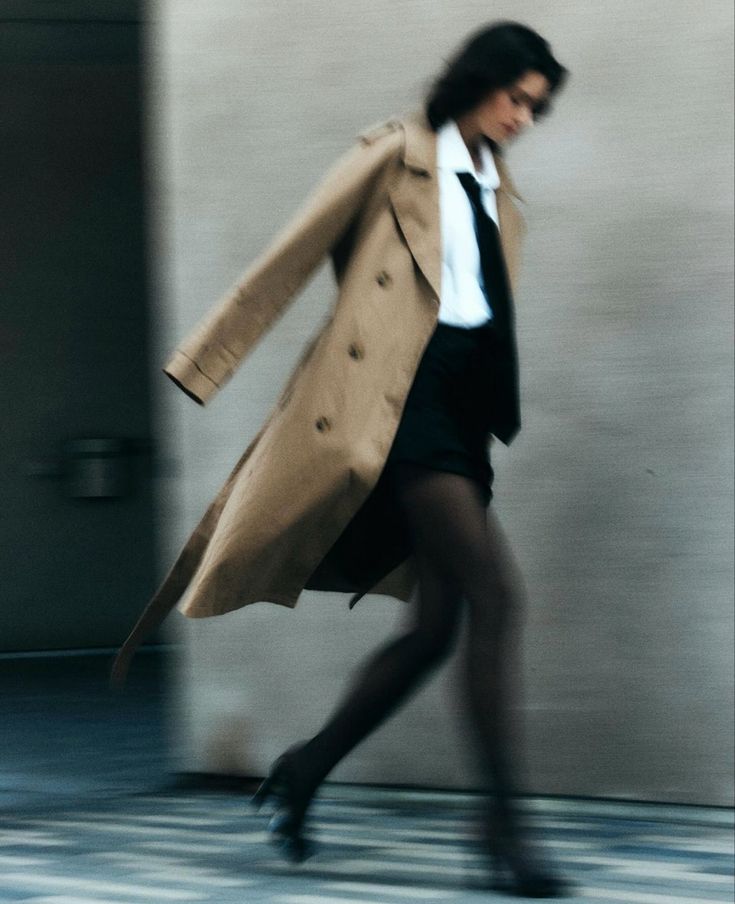 a woman in a trench coat and black skirt is walking down the street with her hand on her hip