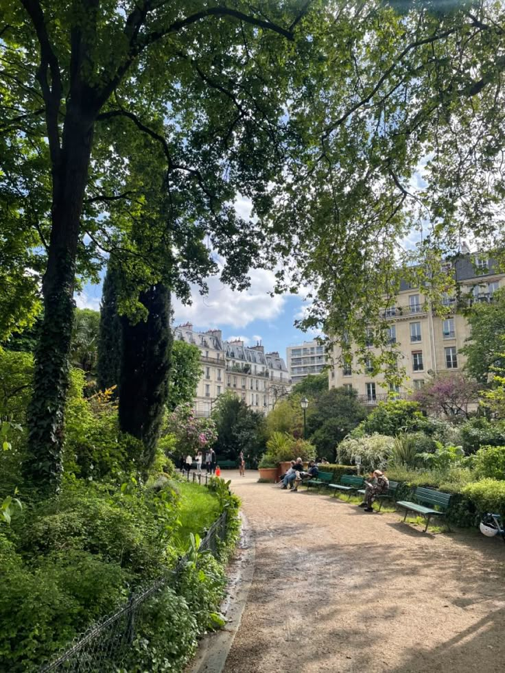 people are sitting on benches in the middle of a park with trees and buildings behind them