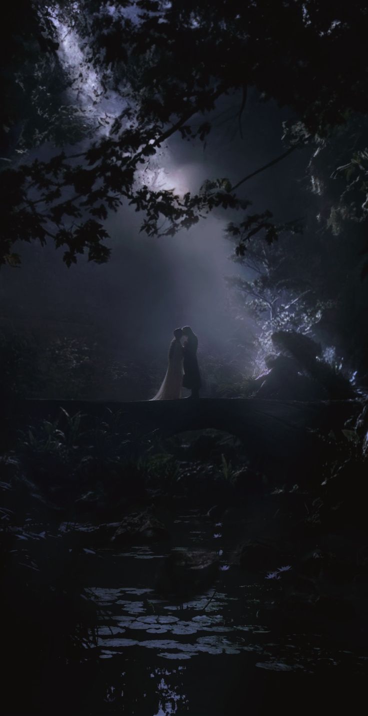 a man and woman are sitting on a bridge in the woods at night with light coming from behind them