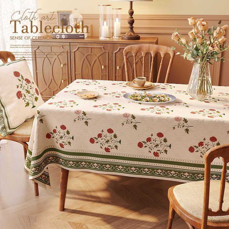 a dining room table covered with a white and green flowered tablecloth next to two chairs