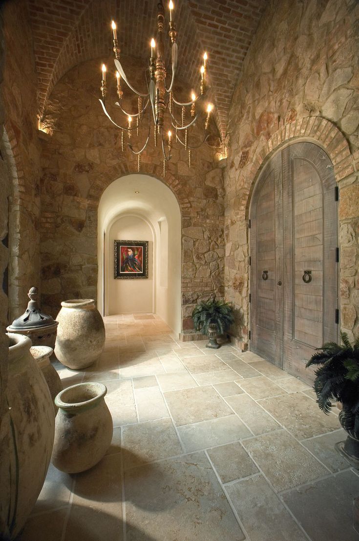 a large hallway with chandelier and potted plants in the center, surrounded by stone walls