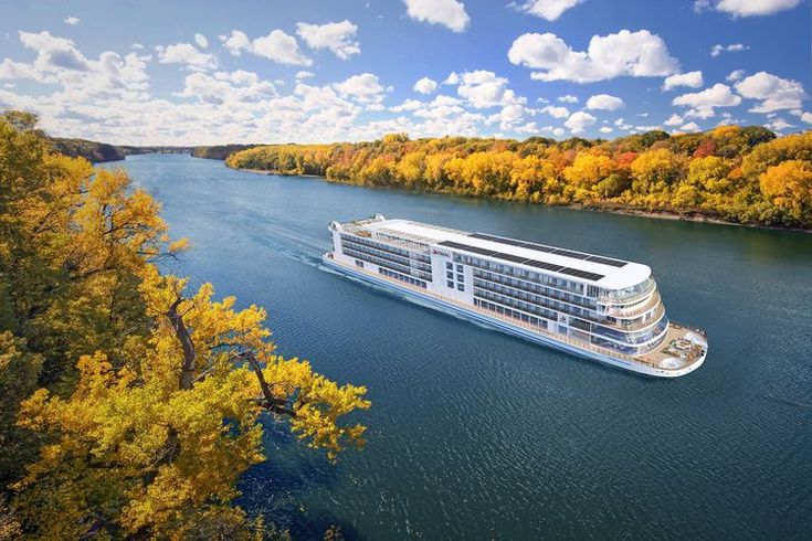 a cruise ship sailing down the river surrounded by fall colored trees and blue sky with white clouds