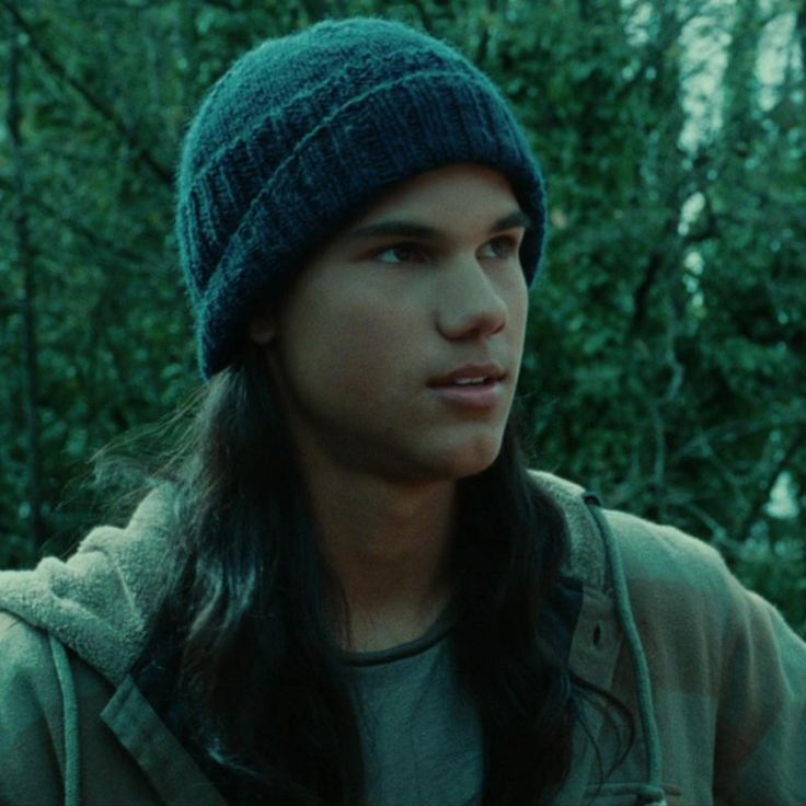 a young man with long hair wearing a beanie in the woods looking to his left