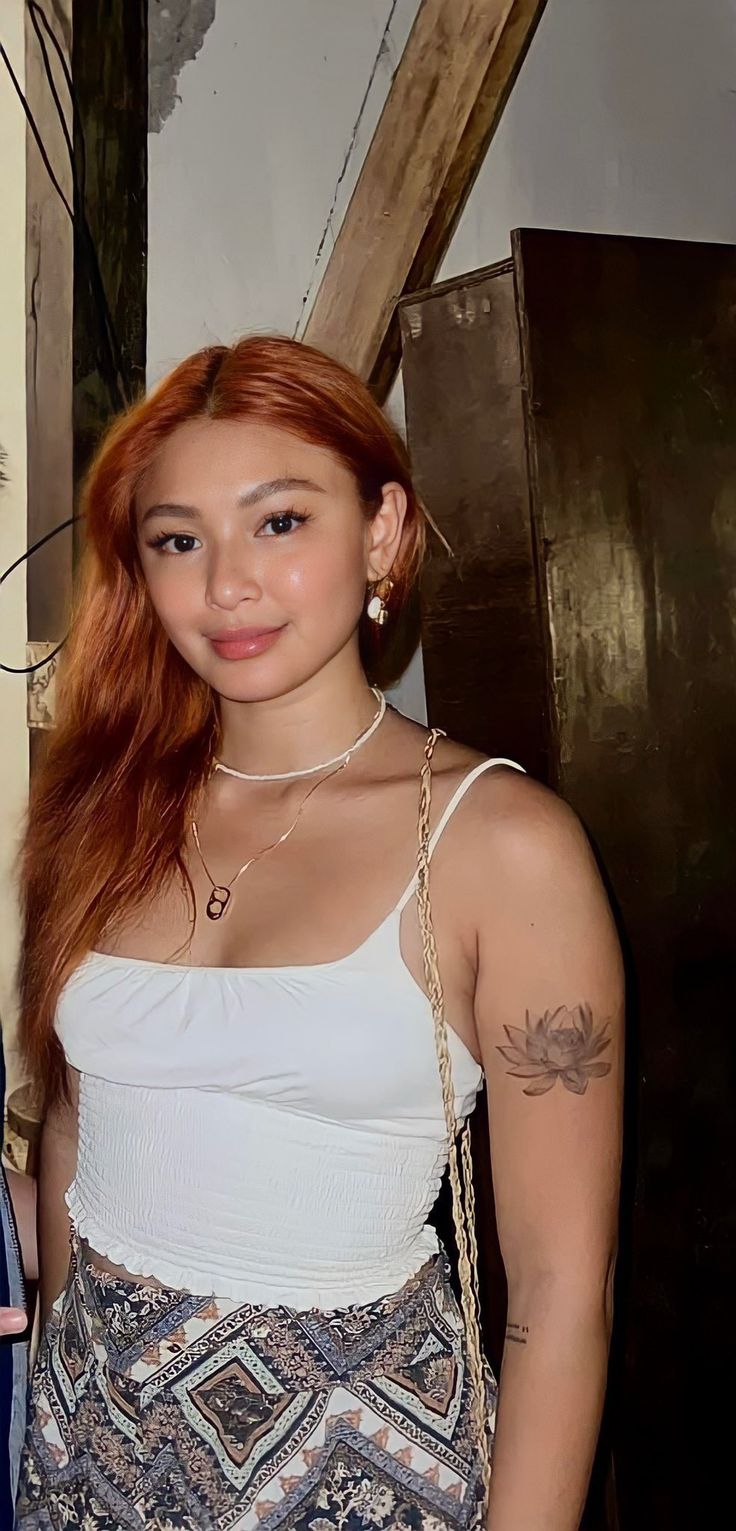 a woman with long red hair standing in front of a wall wearing a white tank top