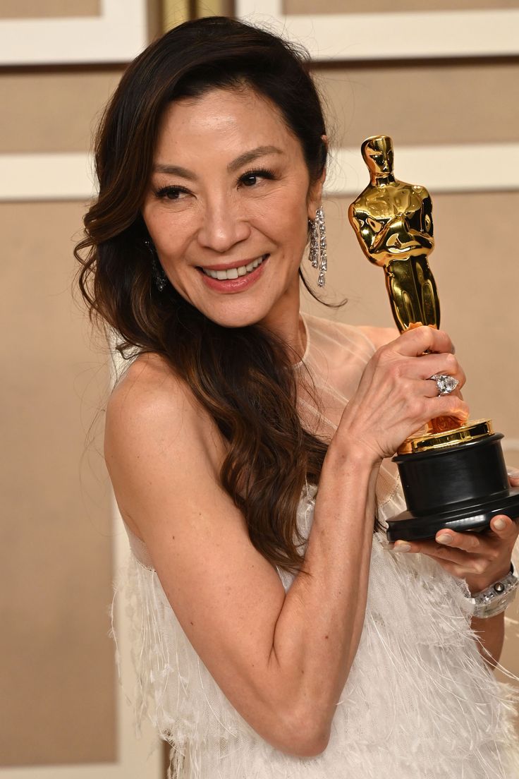 a woman holding an oscar statue in her right hand and wearing a white dress with feathers on it