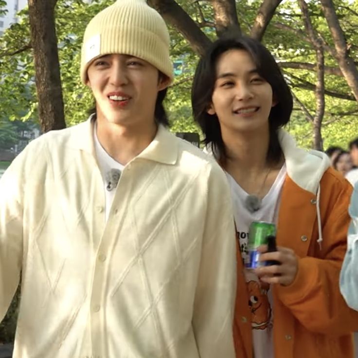 two young men standing next to each other in front of trees and people on the sidewalk