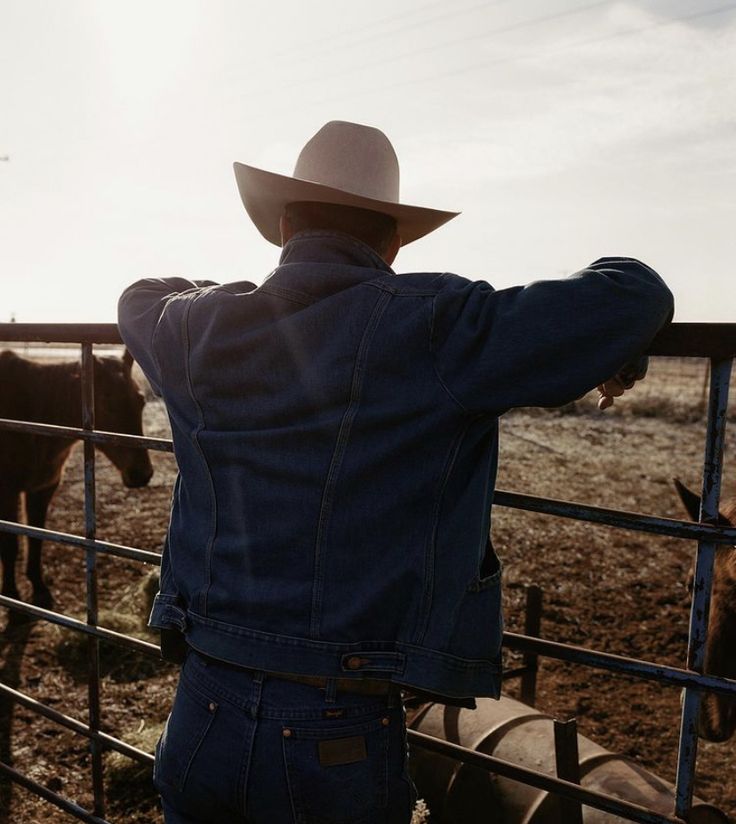 a man wearing a cowboy hat leaning on a fence looking at horses in the field