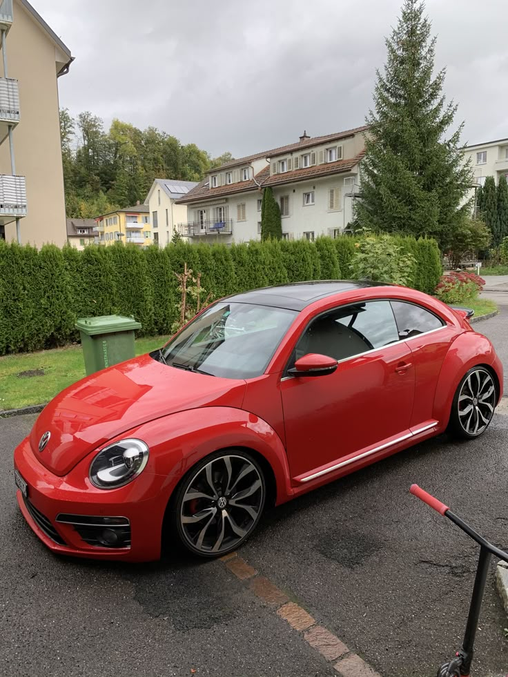 a red volkswagen beetle parked in a parking lot next to some bushes and houses on a cloudy day