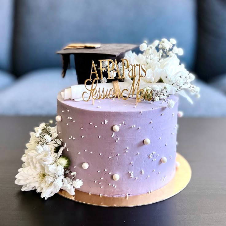 a purple cake with white flowers and a graduation cap on top is sitting on a table