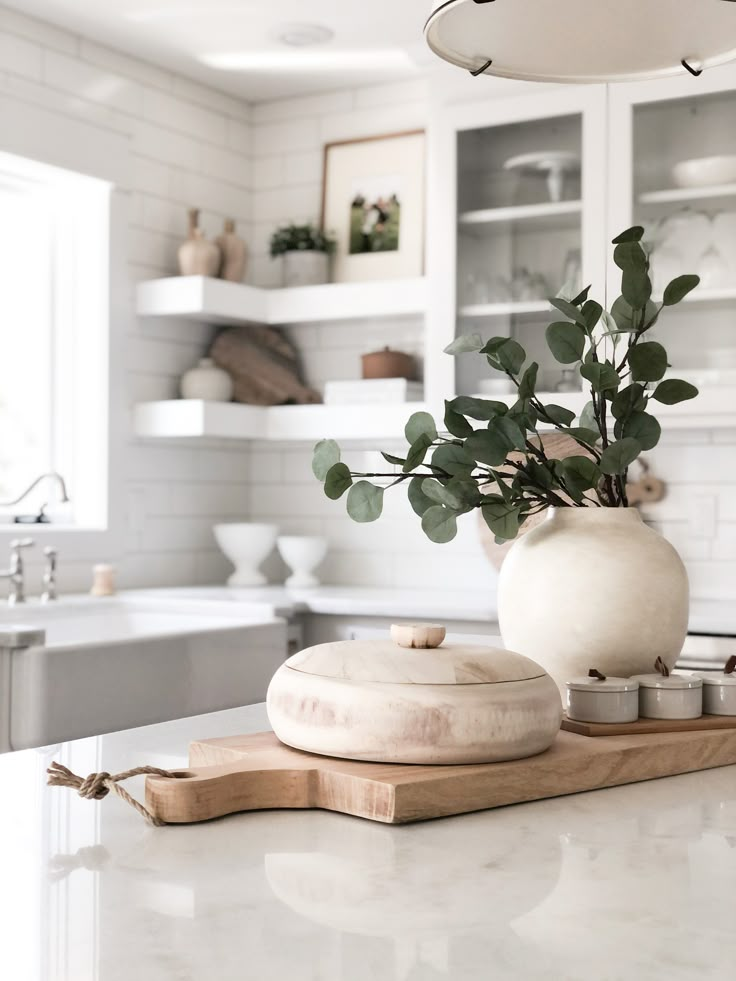 a white vase with some green plants in it sitting on a counter top next to a cutting board