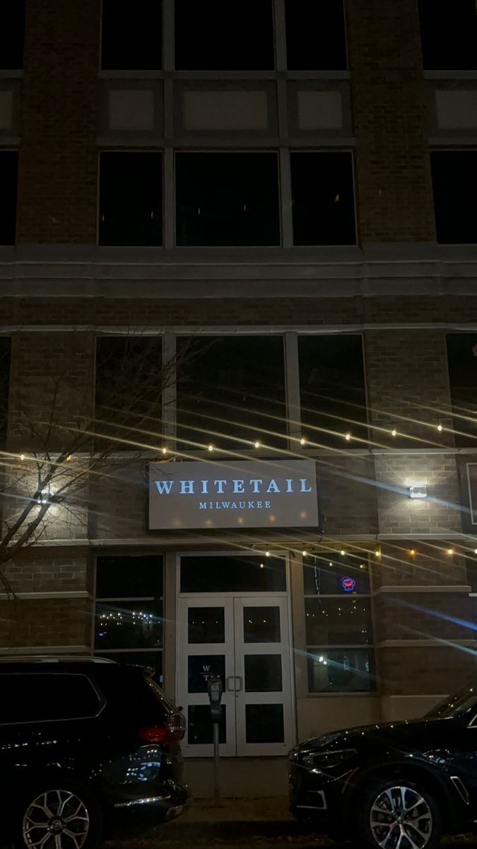 two cars parked in front of a building with lights on the side of it at night