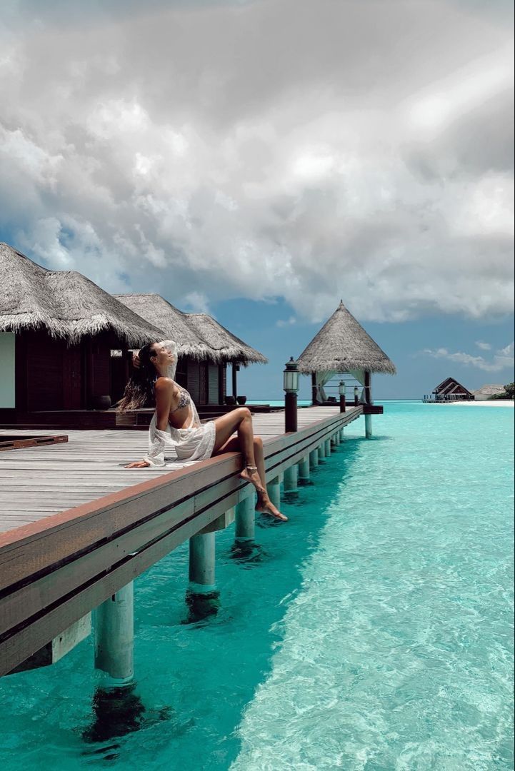 a woman is sitting on a dock in the water near some huts and thatched roofs