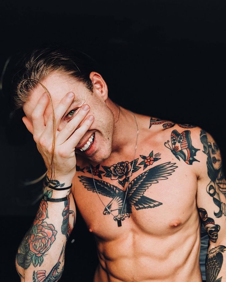 a shirtless man with tattoos holding his hand to his face and looking at the camera