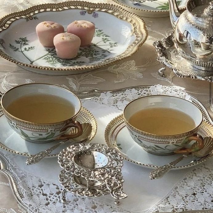 two teacups and saucers on a table with silver dishes, one has candles in it