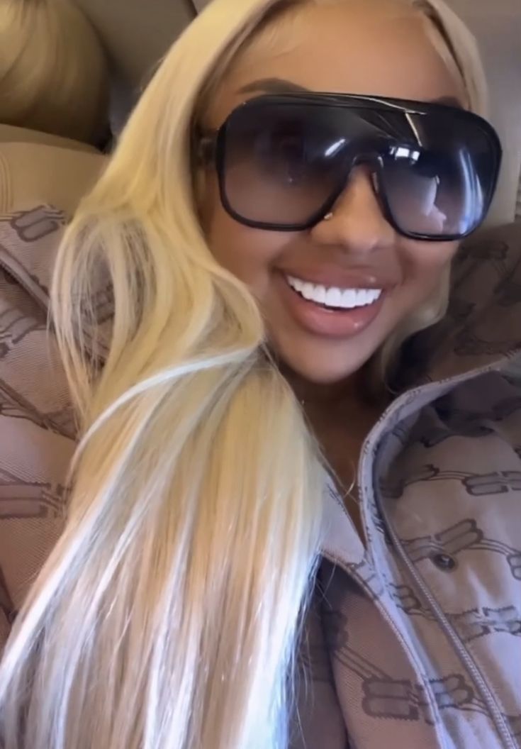 a woman with long blonde hair wearing sunglasses and smiling at the camera while sitting in a car