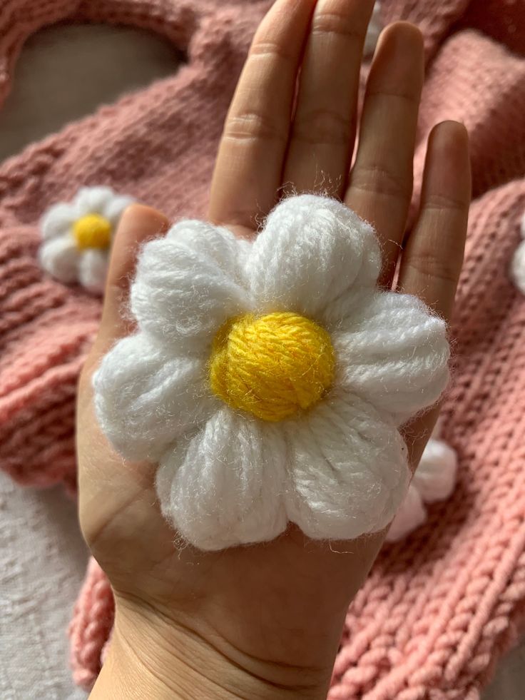 a hand holding a crocheted white and yellow flower on it's palm