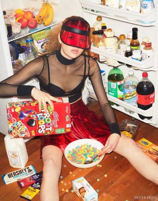 a woman sitting on the floor in front of an open refrigerator holding a bowl of cereal