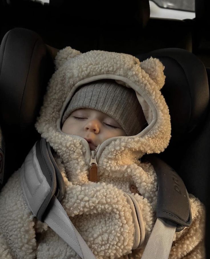 a baby sleeping in a car seat wearing a teddy bear coat and hat with the hood up