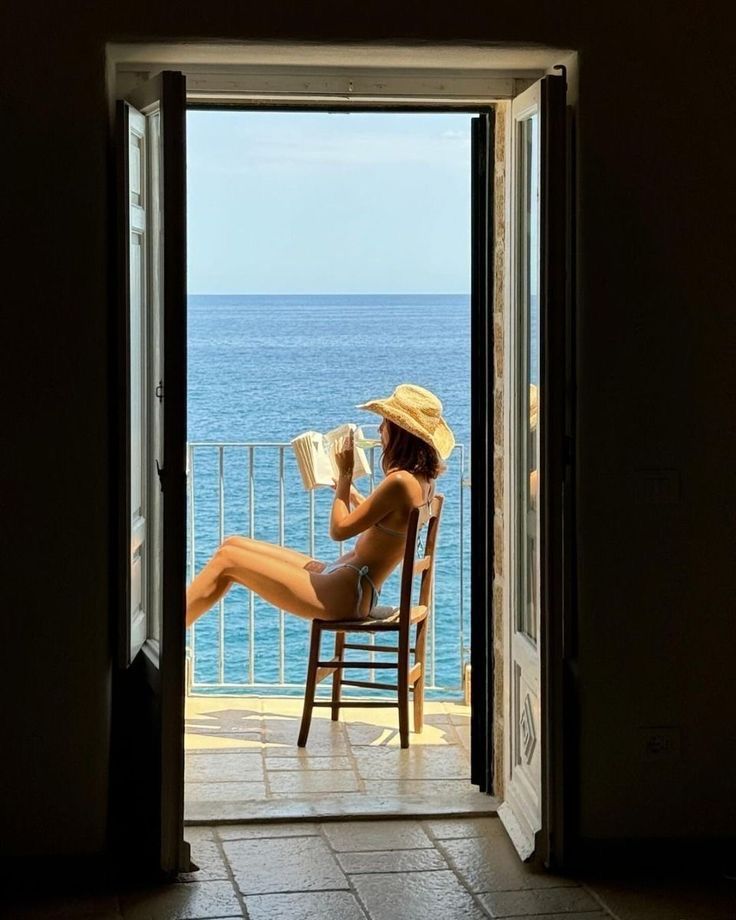 a woman sitting in a chair reading a book while looking out an open door at the ocean