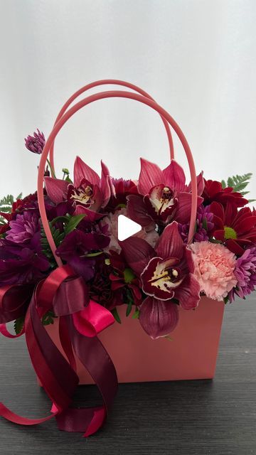 a pink flower basket with purple flowers and red ribbons on the handle is sitting on a table