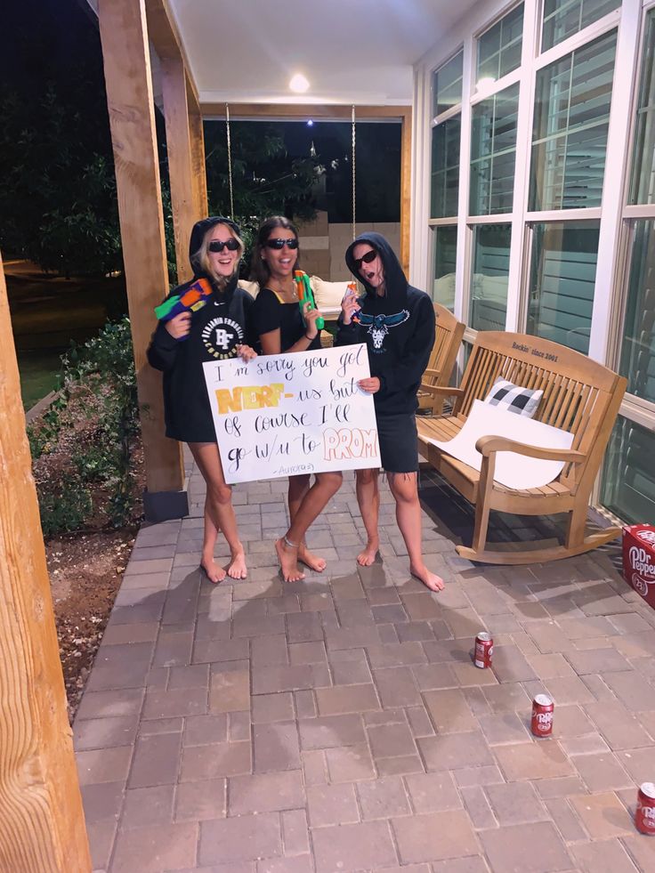 three young women holding a sign in front of a porch with two chairs and one bench