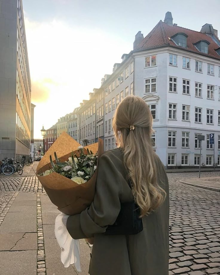 a woman is walking down the street with flowers in her hand and buildings behind her