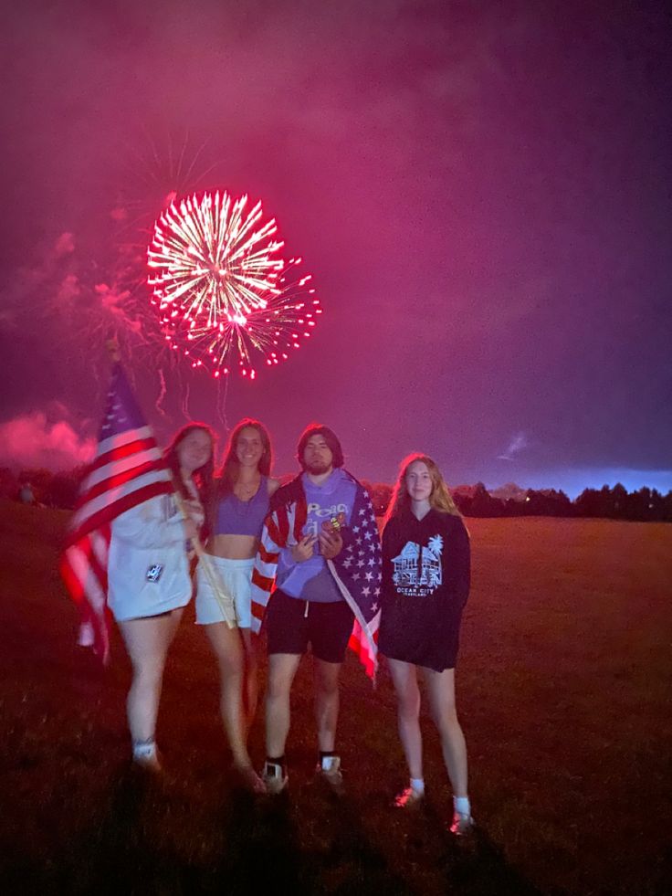 three girls standing in front of fireworks and an american flag on the grass with their arms around each other