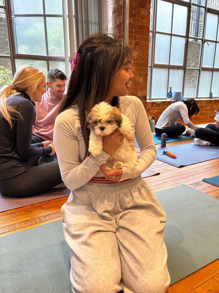 a woman holding a small dog while sitting on a yoga mat in a room full of people