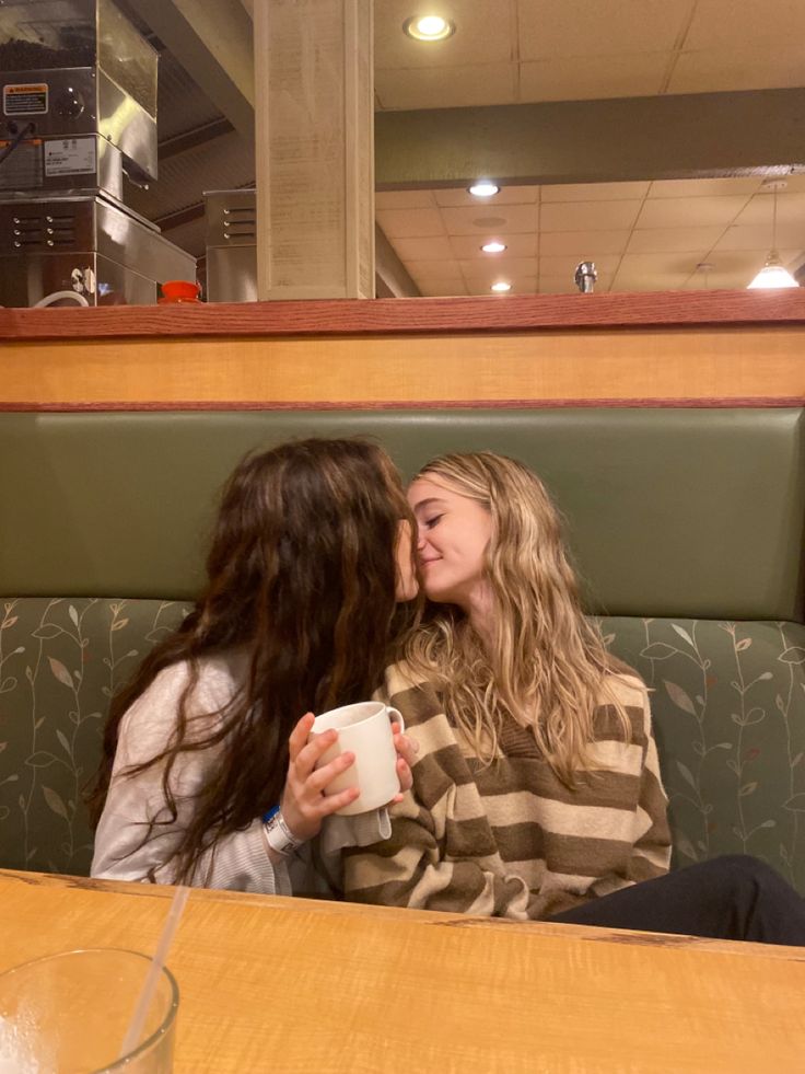 two women sitting at a table sharing a cup of coffee and kissing each other's foreheads