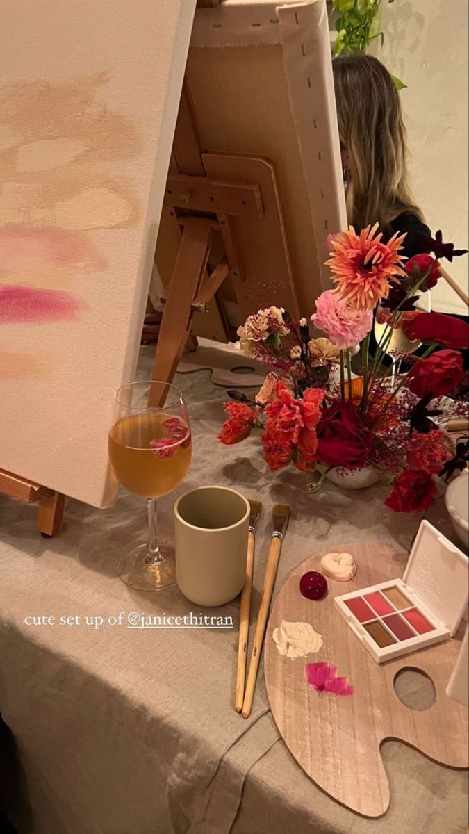 an easel, paintbrushes and flowers on a table with a woman in the background