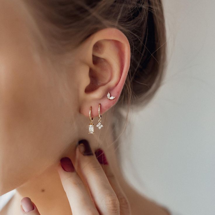 This stacking set features 3 different earrings, all made from 18 karat gold-plated sterling silver. It has: ✅ One small huggie hoop earring with a single, clear-coloured, Baguette-cut zircon stone in a four-prong setting hanging from its lower edge;  ✅ One small huggie hoop earring with a small flower-shaped pendant, which is decorated with four round, clear-coloured zircon stones, and  ✅ One small stud earring with two clear-coloured, oval-shaped zircon stones arranged in a V-shape, with a but Dainty Hoop Earrings, Stacked Earrings, Ear Stack, Hoop Earring Sets, Small Earrings Studs, Delicate Earrings, Baguette Cut, Single Earring, Jewelry Inspo