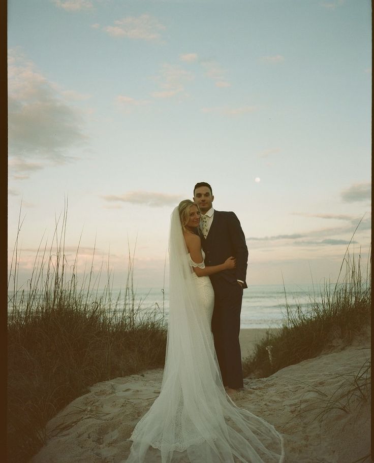 a bride and groom standing on the beach at sunset in front of the sea oats