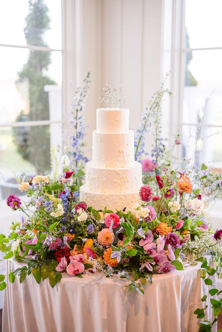 a wedding cake sitting on top of a table covered in flowers and greenery next to a window