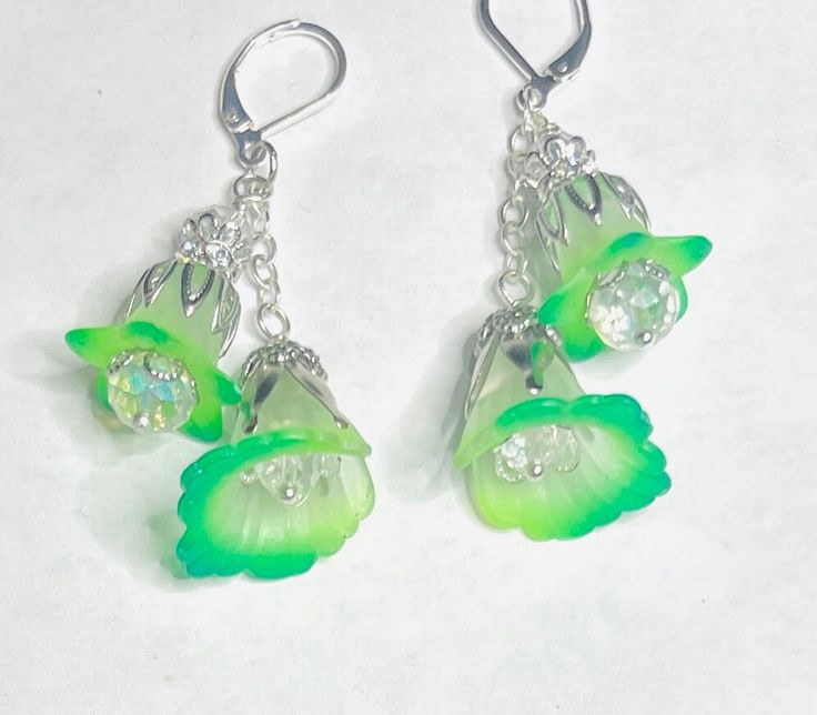 a bold and extraordinary fusion of fluorescent green hues and whimsical fairy vibes. These earrings radiate with vibrant, eye-catching colors, showcasing hand-painted petals that seem to dance with electric energy. Inspired by the captivating glow of fairies, they evoke a sense of wonder and enchantment, making a statement that is both daring and magical. At the heart of these earrings are delicate white flowers, adorned with hand-painted petals in captivating shades of florescent and phthelo green. Each petal is a work of art, showcasing intricate details and a mesmerizing color palette that will catch the eye of everyone around you. Inside each flower, a sparkling clear crystal resides, adding a touch of brilliance and enhancing the overall glamour of the design. The crystals catch the l Whimsical Green Hand Painted Earrings, Green Fairy Earrings For Gift, Fairy Style Green Earrings For Gifts, Green Hand Painted Flower Earrings, Hand Painted Green Flower Earrings, Handmade Green Fantasy Earrings, Bohemian Green Flower Charm Earrings, Handmade Green Whimsical Earrings, Whimsical Green Dangle Earrings