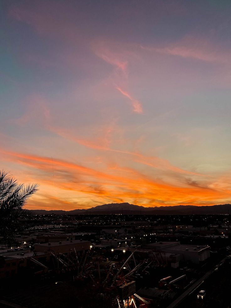 an orange and pink sunset with clouds in the sky above some buildings, trees and mountains