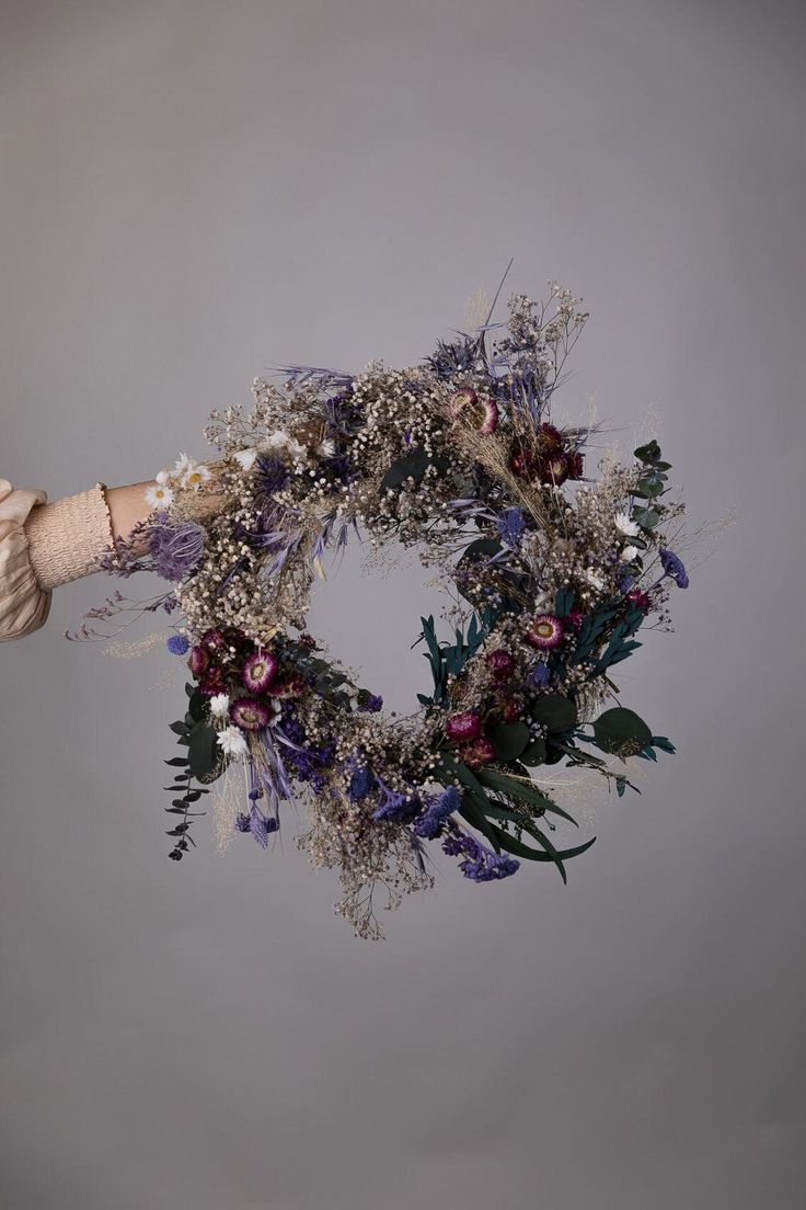 a hand holding a wreath with purple flowers and greenery on the outside, against a gray background