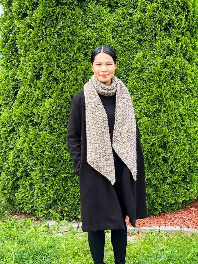 a woman standing in front of a hedge wearing a black coat and gray scarf with her hands on her hips