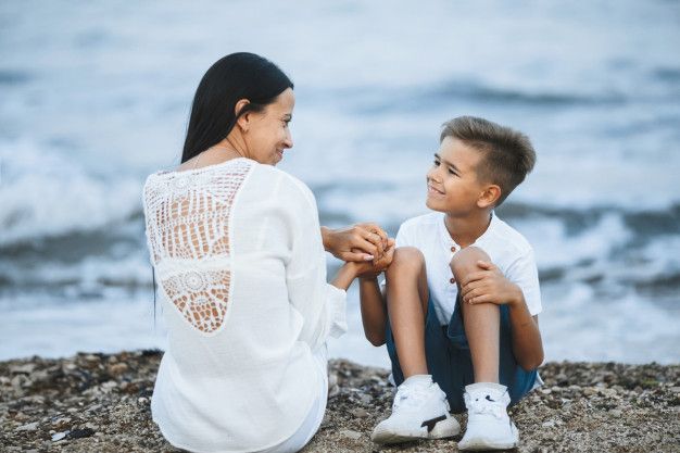 a woman sitting next to a little boy on top of a sandy beach near the ocean