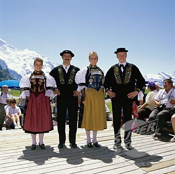 three men and two women dressed in traditional clothing standing on a wooden deck with mountains in the background