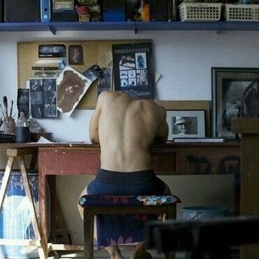 a shirtless man sitting at a desk in front of a painting on the wall