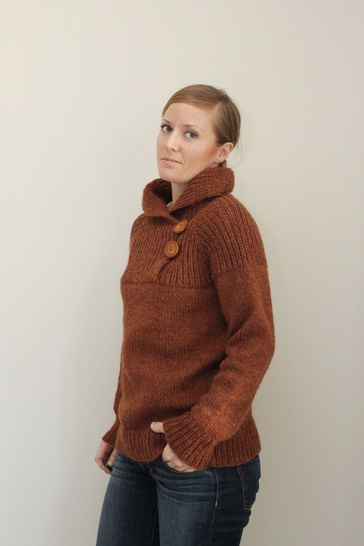 a woman standing in front of a white wall wearing a brown sweater and blue jeans