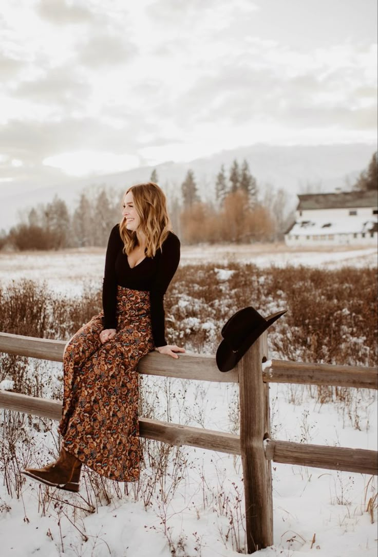 a woman sitting on a fence in the snow wearing a dress and black cowboy hat